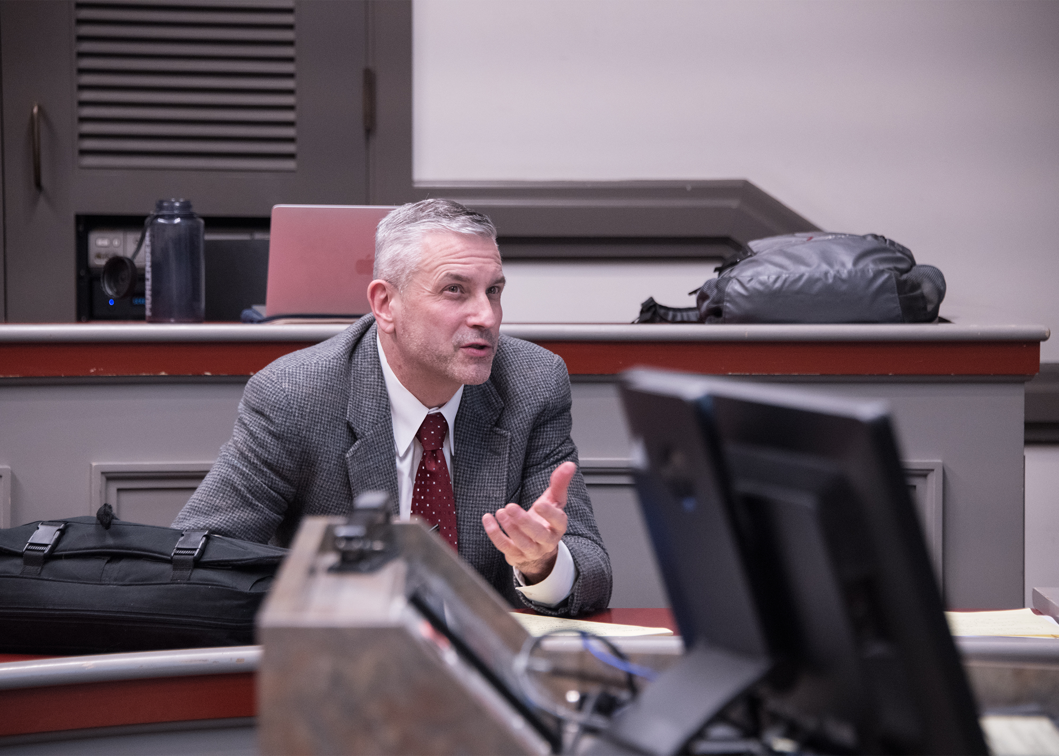 A person in a suit and graying hair speaks to someone off-camera in a classroom.